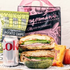 Zingerman's Deli sandwich with pickle, can of soda, bag of potato chips, mini Zzang candy bar, apple, napkins and wet nap in front of red plastic Zingerman's Deli bag.