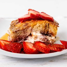 Slice of stuffed challah french toast with preserves and cream cheese, garnished with strawberries and maple syrup.