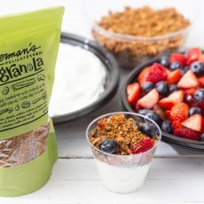 Yogurt, homemade granola, fresh berries behind a yogurt parfait in a clear plastic cup.