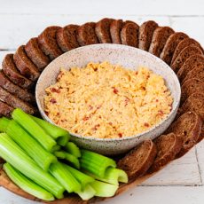 Creamy pimento cheese dip in a speckled bowl, surrounded by sliced pumpernickel bread and fresh celery sticks - a classic appetizer spread.