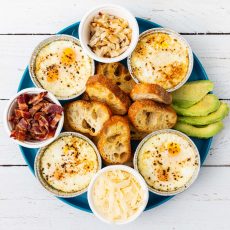 4 individual aluminum ramekins, each containing 2 shirred eggs arranged on a blue plate with ramekins of bacon lardons, roasted onions and Grana Padano cheese. Slices of avocado and toasted baguette slices are also on the plate.