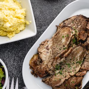 Sliced tender beef brisket arranged on a white serving platter, garnished with fresh herbs, alongside a bowl of creamy mashed potatoes