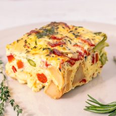 Square slice of frittata with roasted peppers, potatoes, onions and cheddar on an off-white plate, garnished with herbs.