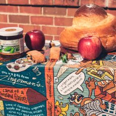 Small loaf of Challah Bread, two apples, two rugelach and a jar of honey on top of a cartoon decorated gift box in front of a brick wall