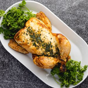Golden roasted whole chicken topped with fresh herbs and lemon zest, presented on a white platter with sprigs of curly parsley