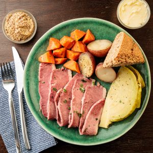 An overhead shot of a green ceramic plate featuring sliced corned beef garnished with fresh herbs, a braised cabbage wedge, halved red potatoes, and chunky roasted carrots. A triangle of seeded soda bread rests on the right side of the plate. Small glass bowls of whole grain mustard and butter are visible in the upper corners. Silverware and a blue polka-dot napkin are arranged to the left on a dark wood surface.