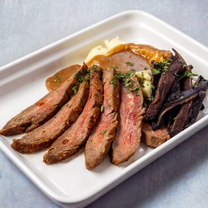 Medium-rare flank steak sliced against the grain, served with buttery mashed potatoes, rich gravy, and grilled portobello mushrooms on a white rectangular plate